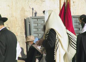 Person im Gebetsgewandl an der Klagemauer mit anderen Personen in der Nähe, eine sitzt mit einem beigen Hut. Roter Stoff im Hintergrund.