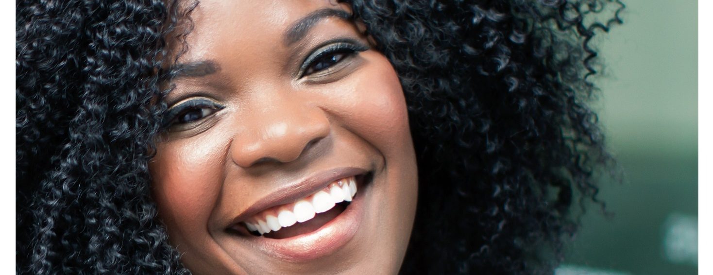 Eine Nahaufnahme einer Frau mit lockigem Haar, die strahlend lächelt, deren weiße Zähne sichtbar sind und die direkt in die Kamera blickt.