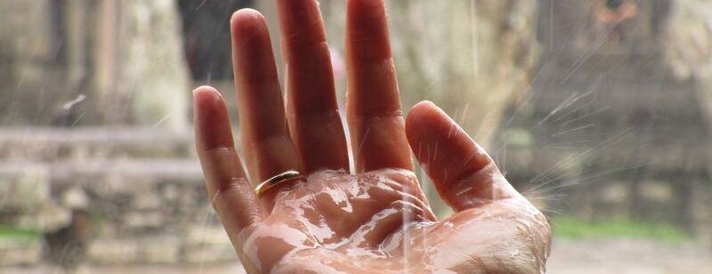 Hand mit Regentropfen, ausgestreckt in einem Regenguss mit verschwommenen Steinstrukturen im Hintergrund.