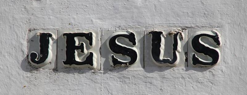 Das Wort „JESUS“ steht in schwarzen Buchstaben auf weißen Fliesen vor einer strukturierten weißen Wand.