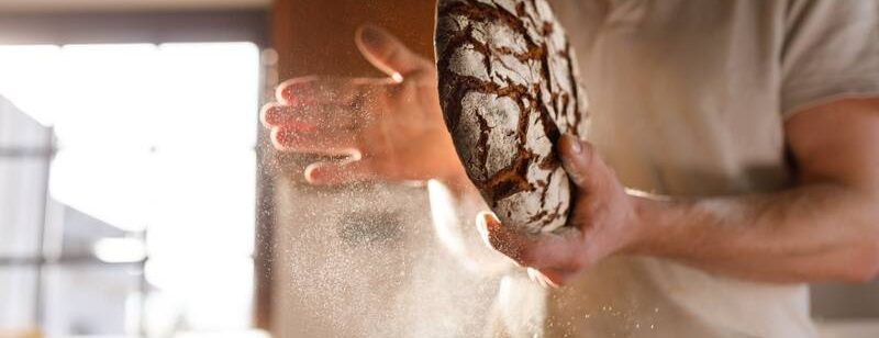 Person hält frisch gebackenes handwerklich hergestelltes Brot in der Hand, Mehlstaub in der Luft, in einer warm beleuchteten Küche.